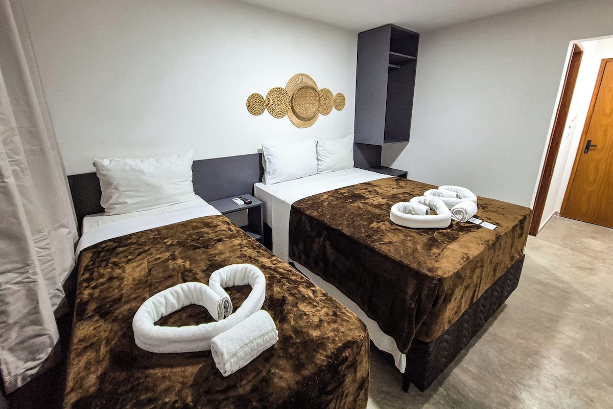 A hotel room with two beds, each with a brown bedspread and white pillows. There are two white towels on the beds. A small table with a phone and a remote control is between the beds.