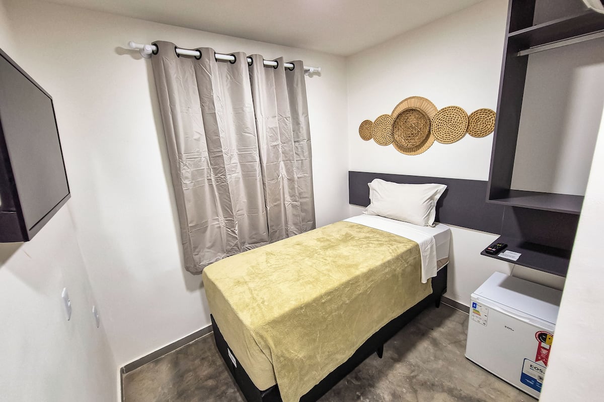 A bedroom with a bed, a small white refrigerator, a wall-mounted shelf, and a curtain.