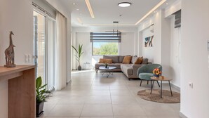 A spacious living room with a sofa, a coffee table, and a chair. There is a large window providing natural light and a view of greenery outside. - AeNaos Luxury Villa (Chania)