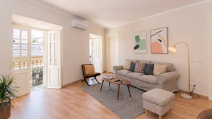 A spacious living room with a sofa, a coffee table, and a chair. There are two doors leading to a balcony, and a potted plant is visible. - Casa Telhados | Historic Center | Private Terrace (Faro)
