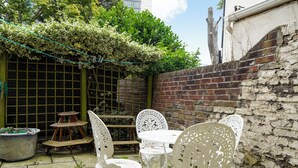 A patio area with white metal chairs and a small table, surrounded by a brick wall and greenery. - The Earl's Court Wonder - Adorable 1bdr Flat With Terrace (London)