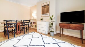 A dining area with a table and chairs, a television on a stand, a potted plant, and a wall-mounted lamp. - The Earl's Court Wonder - Adorable 1bdr Flat With Terrace (London)