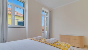 A hotel room with a bed, a bedside table, a window with curtains, and a view of buildings outside. - Funchal City Nest III (Funchal)