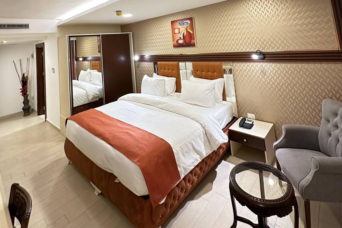 A hotel room with a large bed, a bedside table with a phone, a chair, a small table, and a mirror.
