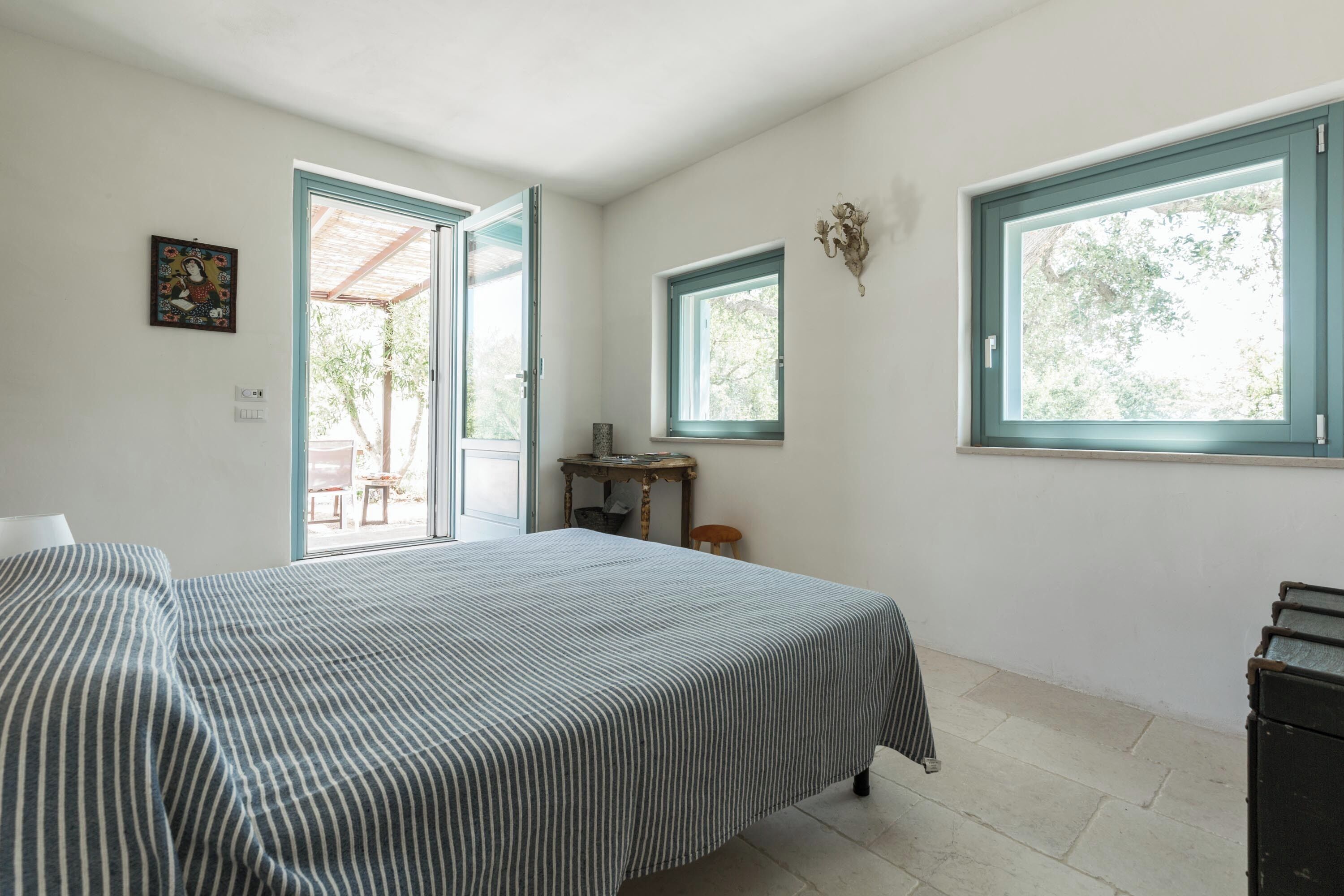 A bedroom with a bed, two windows, a door leading outside, and a small table with a chair.
