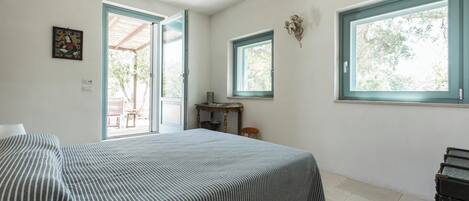A bedroom with a bed, two windows, a door leading outside, and a small table with a chair.