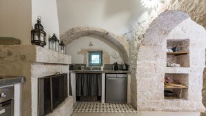 Une cuisine avec des murs en pierre, un sol carrelé, un four encastré, un lave-vaisselle et une fenêtre avec des rideaux.