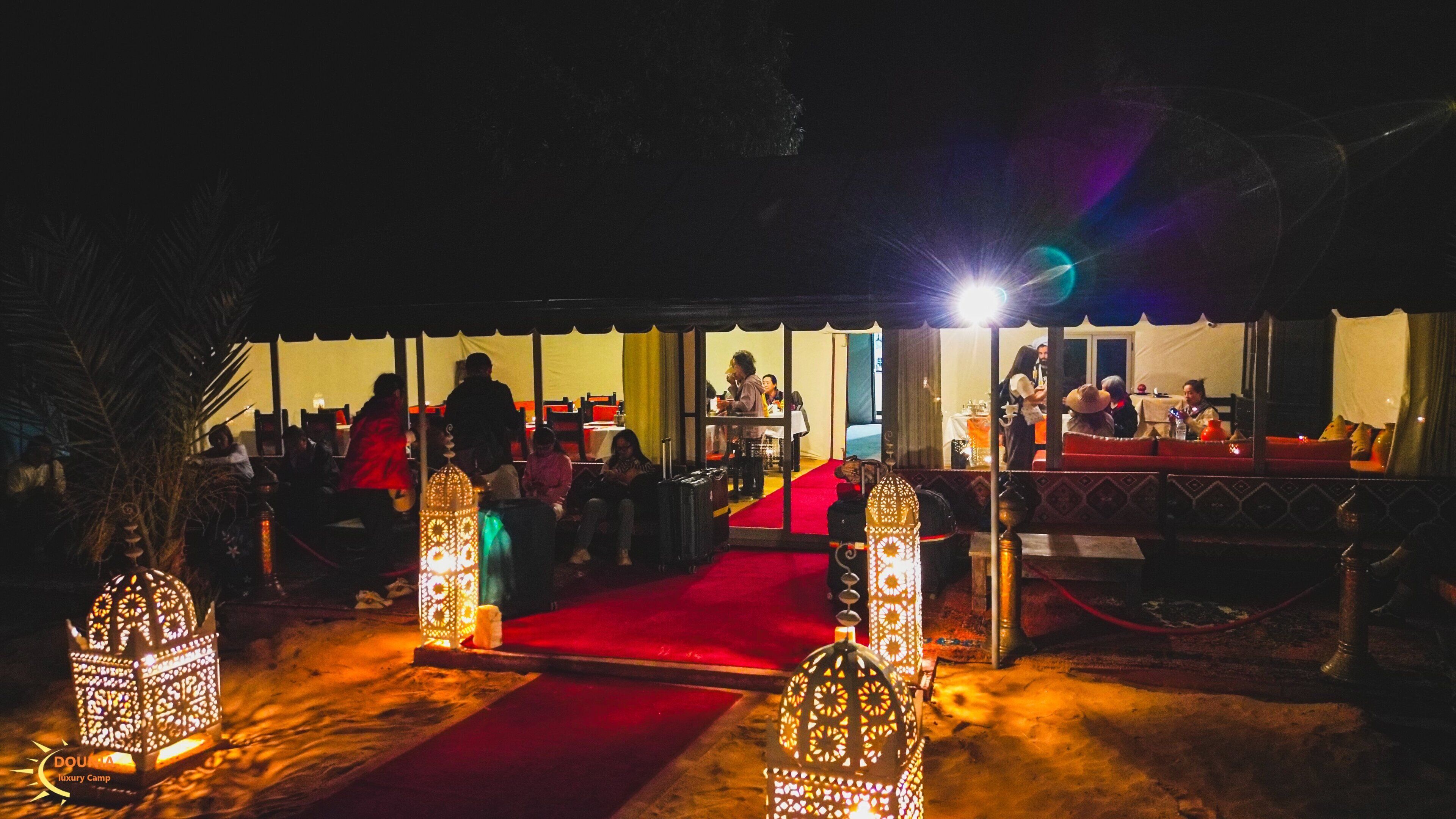 Un espacio de comedor al aire libre con alfombra roja, faroles ornamentales y un área de estar cubierta.