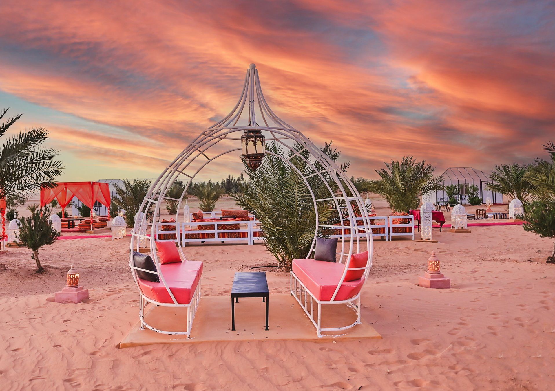 A desert setting with a white gazebo structure, red cushioned seating, and a small table.