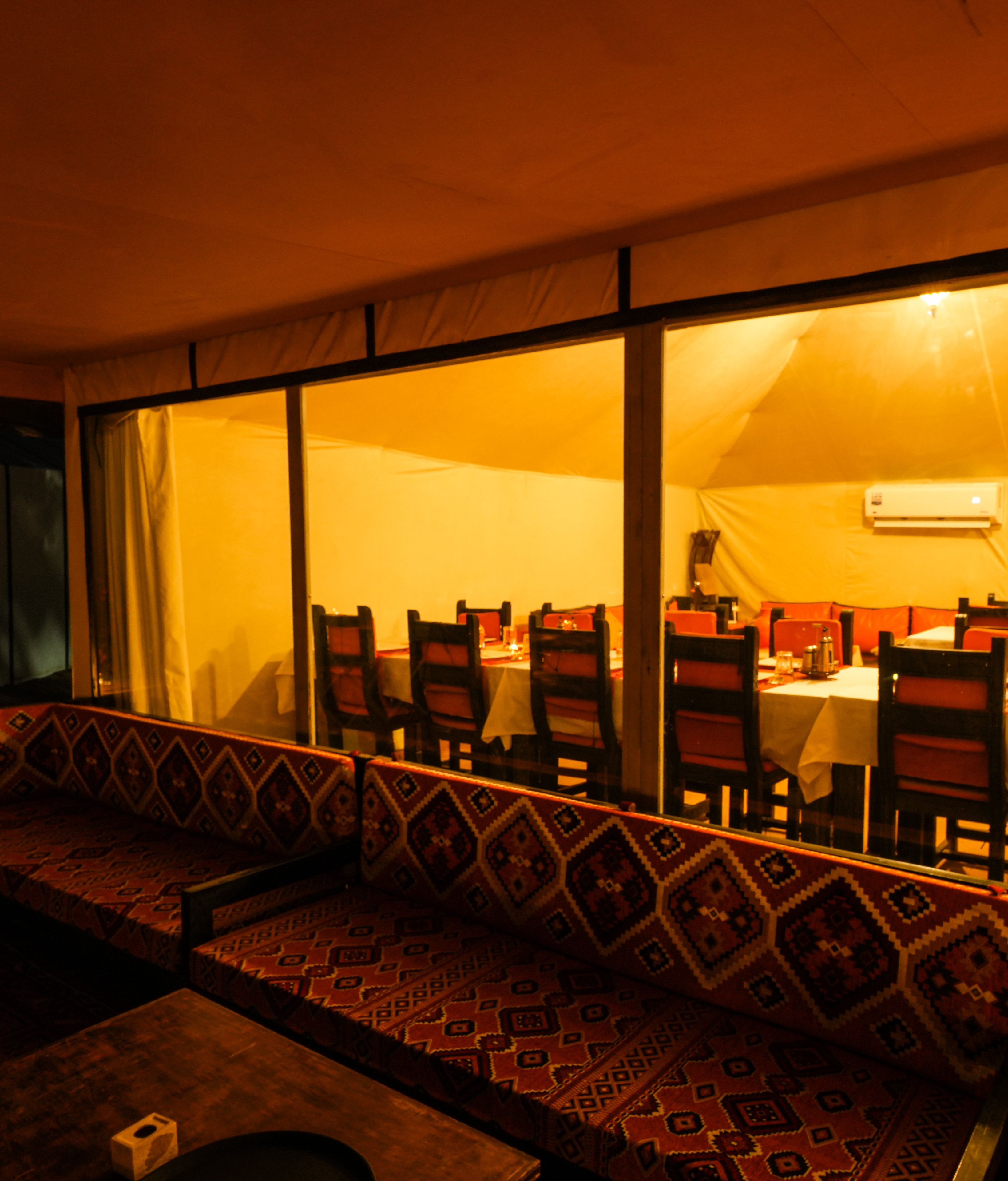Un área de comedor interior con una mesa larga y sillas, un banco con cojines estampados y una ventana con cortinas.
