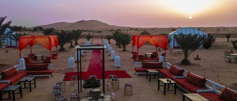 A desert camp with a central red carpet leading to a seating area with red cushions and white lanterns.