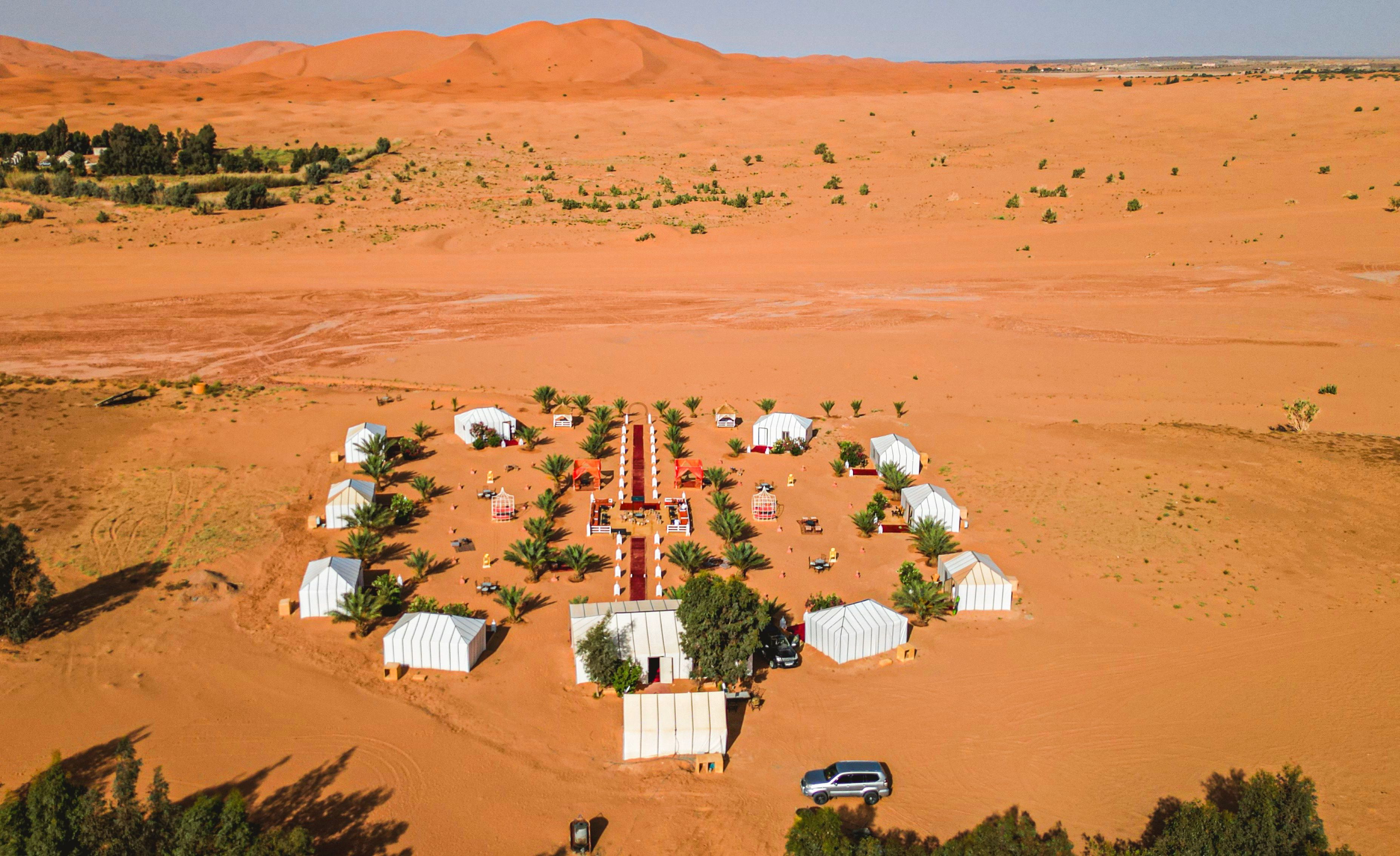 An aerial view of a desert camp with several white tents arranged in a circular pattern around a central structure.
