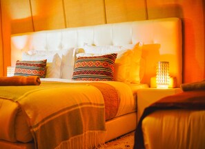 A hotel room with a large bed, multiple pillows, a bedside table with a lamp, and a blanket. - Dounia Luxury Camp (Rissani)