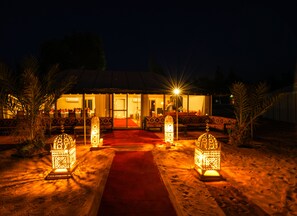 A building with a red carpet leading to the entrance, surrounded by palm trees and illuminated by lanterns. - Dounia Luxury Camp (Rissani)