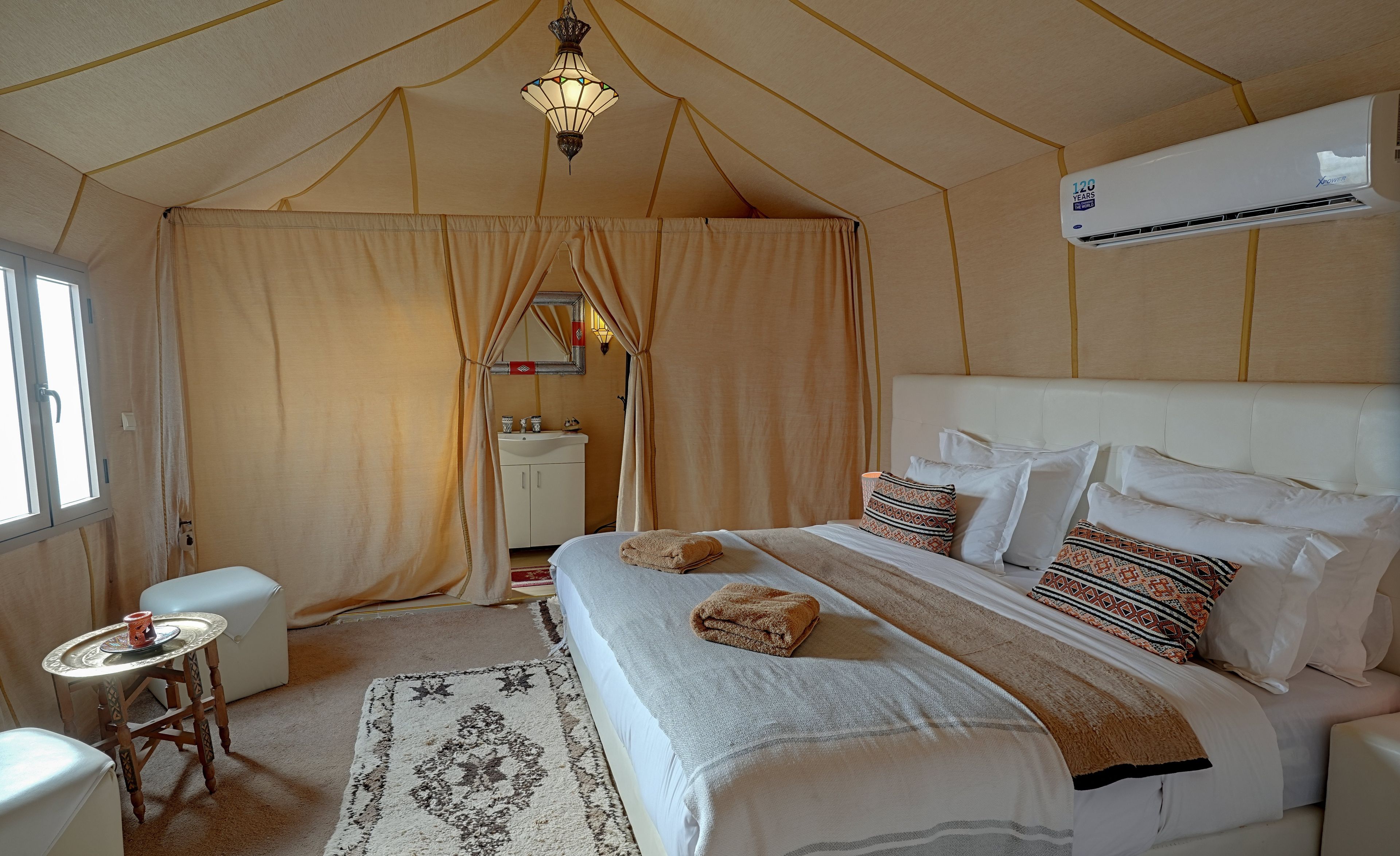 A tent-style room with a bed, a small table, a chair, a sink, and a window.