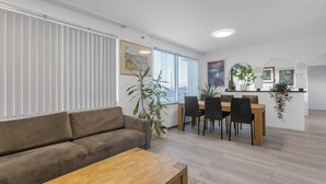 A modern living room with a sofa, coffee table, dining area with chairs, and a kitchen counter with plants.