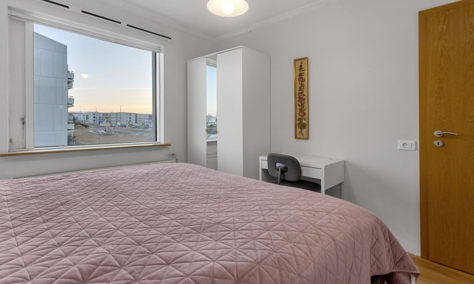 Um quarto com uma cama grande, uma escrivaninha, uma cadeira, um guarda-roupa e uma janela com vista para a cidade.