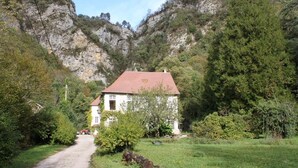 Ein weißes Haus mit rotem Dach, umgeben von Grün und einem felsigen Berg im Hintergrund.