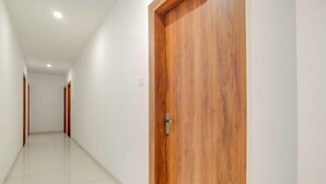 A hallway with a wooden door on the right, white walls, and a tiled floor. - Hotel Apple  (Indore)