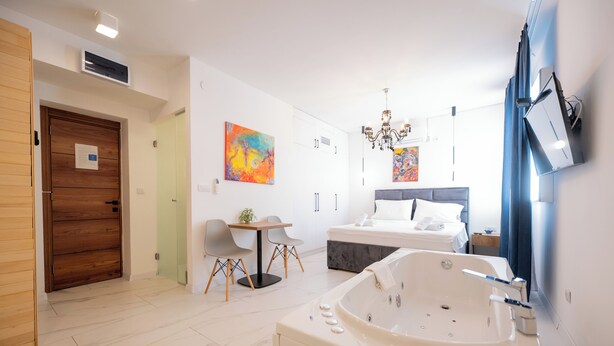 Una camera d'albergo moderna con una vasca da bagno grande, un letto con testiera grigia, un tavolino con sedie e un quadro astratto dai colori vivaci appeso al muro.