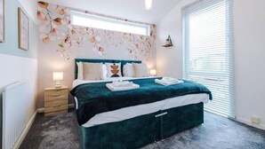 A bedroom with a large bed, a headboard with a floral pattern, a bedside table with a lamp, and a window with blinds.