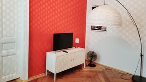 A room with a red wall, a white door, a white cabinet with a television, a floor lamp, and a standing fan. - Wipplinger Home Apartment Vienna (Vienna)