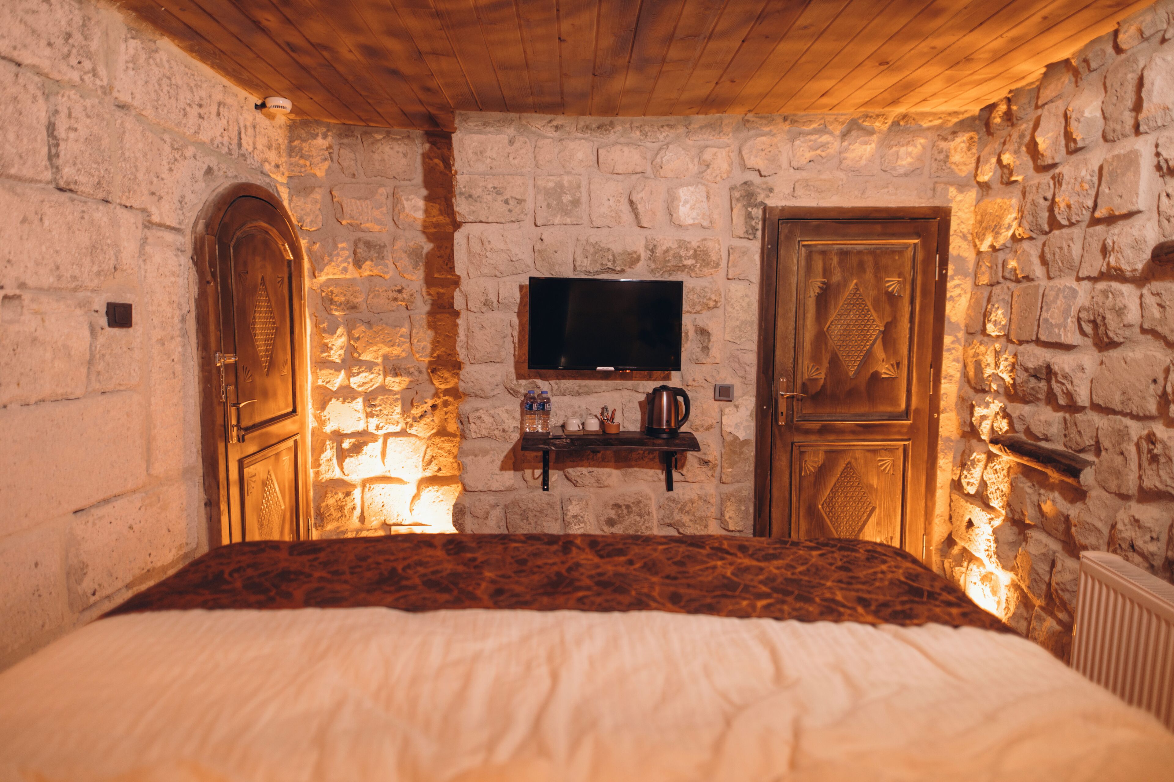 A stone-walled room with a bed, a television mounted on the wall, and a wooden door.