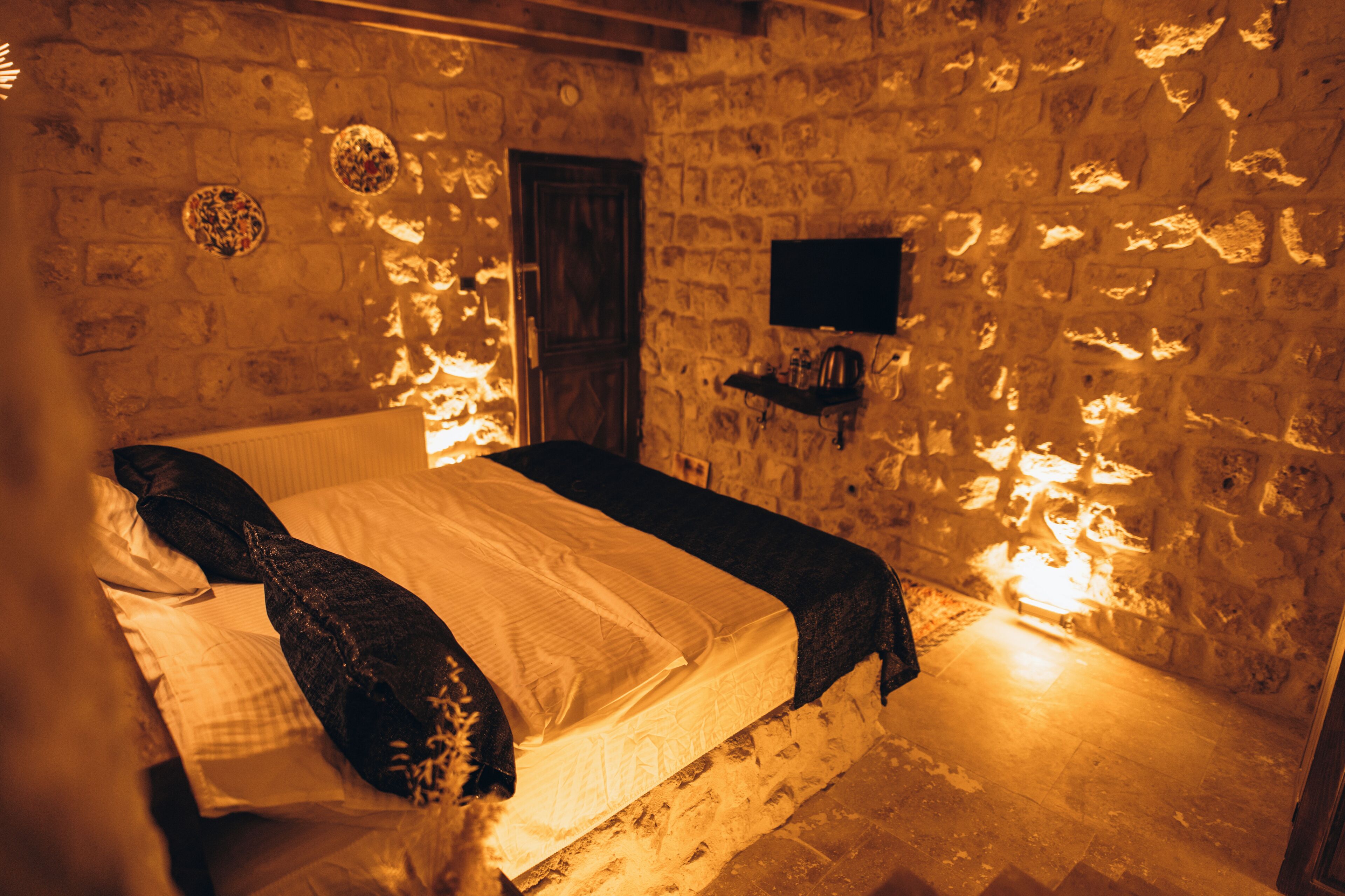 Una habitación con paredes de piedra, una cama, un televisor en la pared y una puerta.