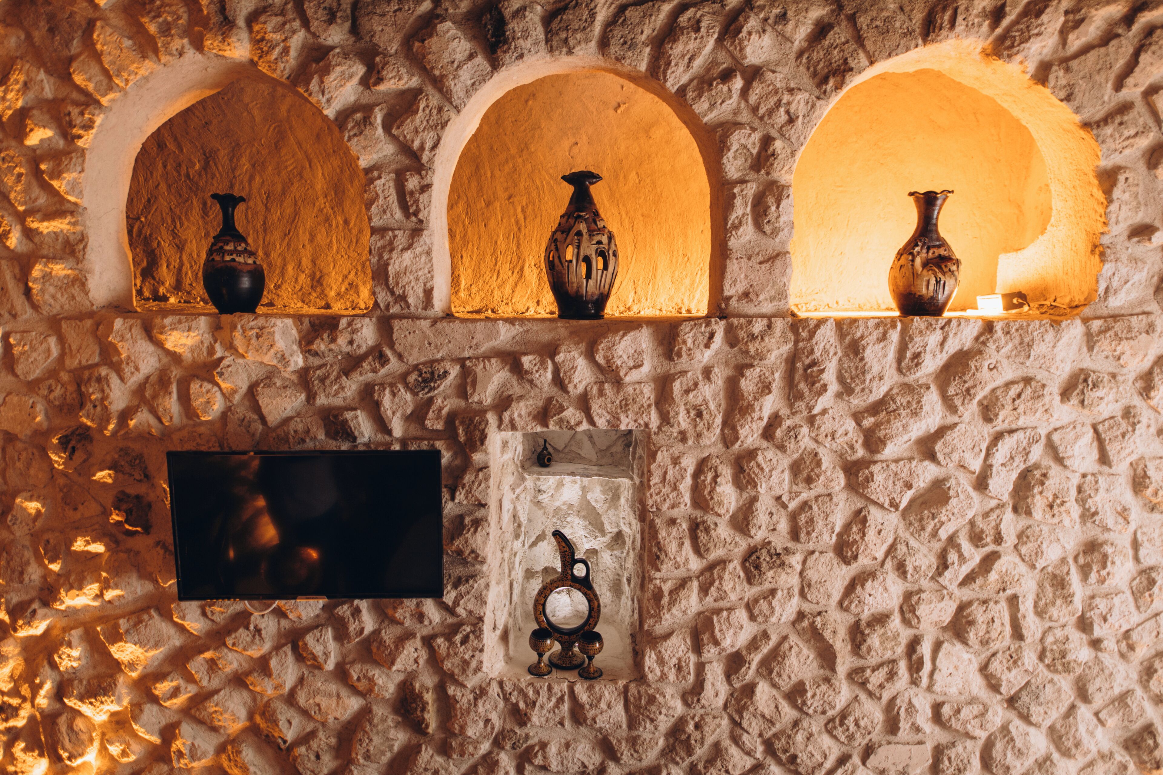 A stone wall with three arched niches, each holding a vase. Below, a small framed picture and a black rectangular object are mounted.