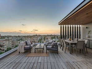 A rooftop terrace with a wooden deck, outdoor seating, and a view of the cityscape during sunset. - Sanders Olive Shared Pool Apartments (Paphos)