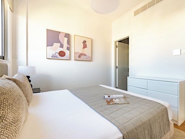 A modern bedroom with a large bed, a bedside table with a lamp, a dresser, and two abstract paintings on the wall. - Sanders Olive Shared Pool Apartments (Paphos)
