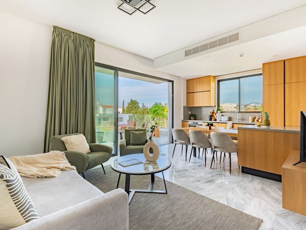 A modern living room with a sofa, chairs, a coffee table, and a view of the city through large windows. - Sanders Olive Shared Pool Apartments (Paphos)