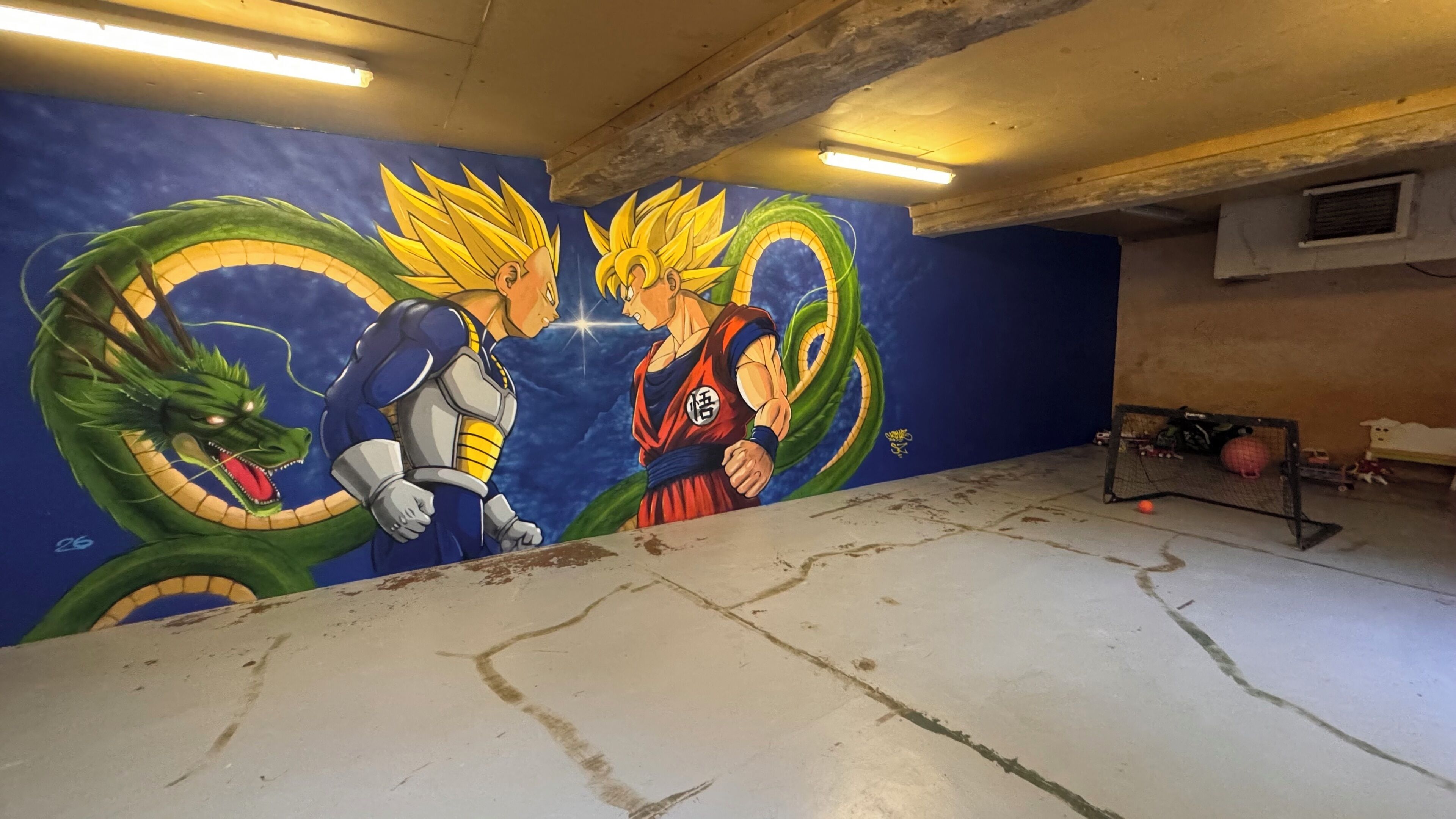 Une pièce décorée d’une fresque représentant deux personnages de Dragon Ball Z, avec un sol en béton et une petite étagère sur laquelle sont disposés divers objets.