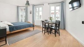 A bedroom with a bed, bedside table, and a desk with a chair. There is a window with curtains, a TV mounted on the wall, and a view of buildings outside.