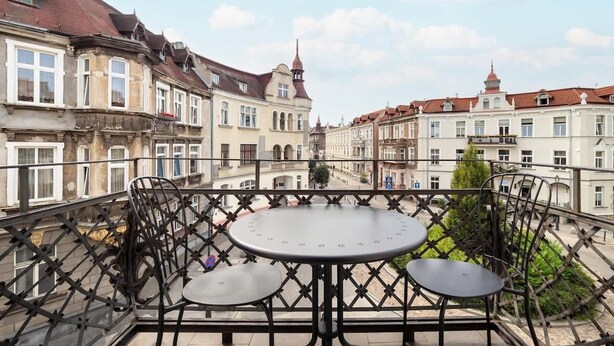 A balcony with a round table and four chairs, overlooking a street with historic buildings and a church steeple.