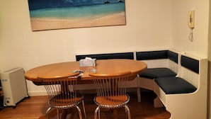 A dining area with a round table, two chairs, a bench with cushions, a painting of a beach, and a telephone on the wall.