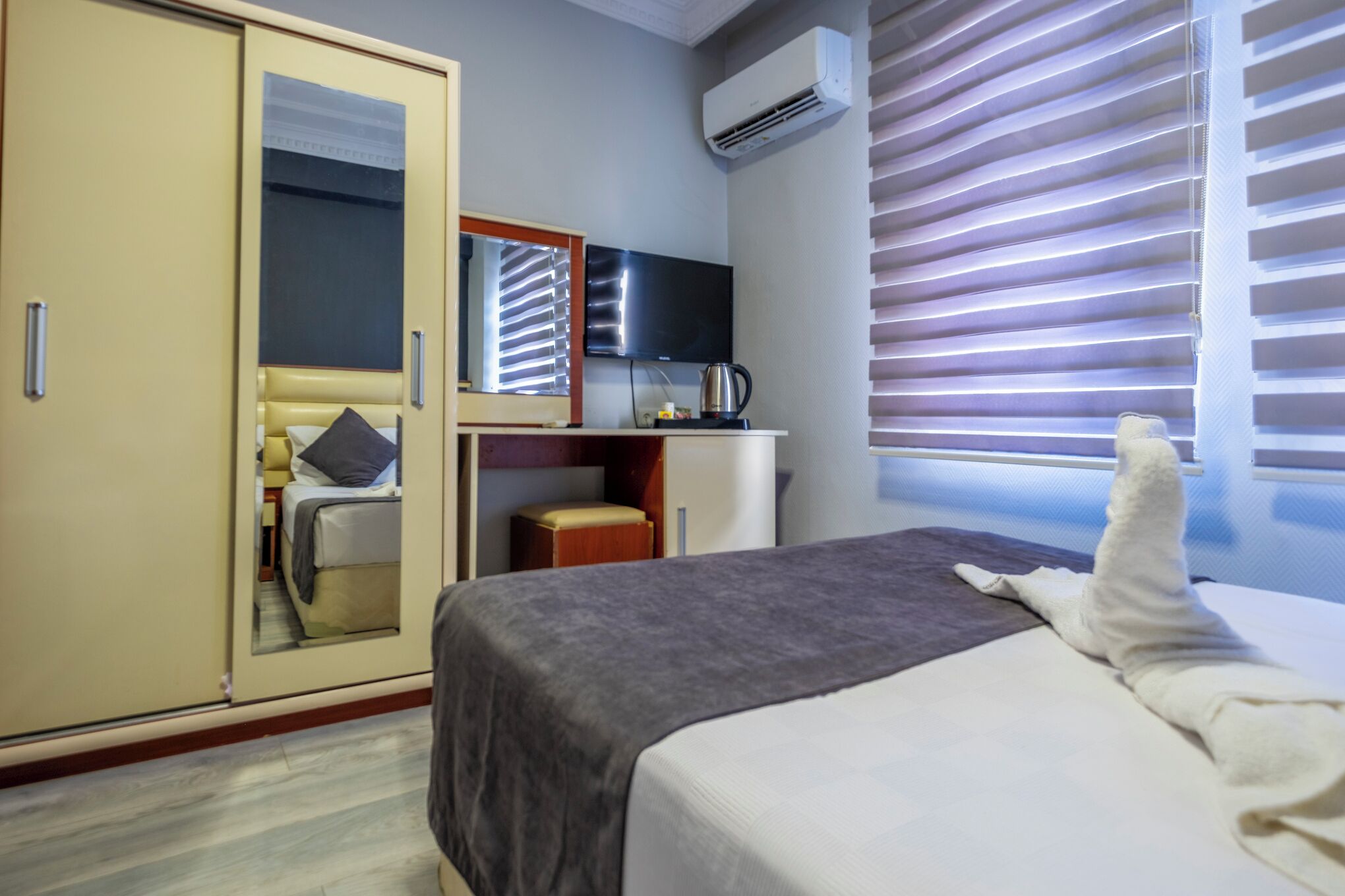 A hotel room with a bed, a wardrobe, a mirror, a television, and a window with blinds.
