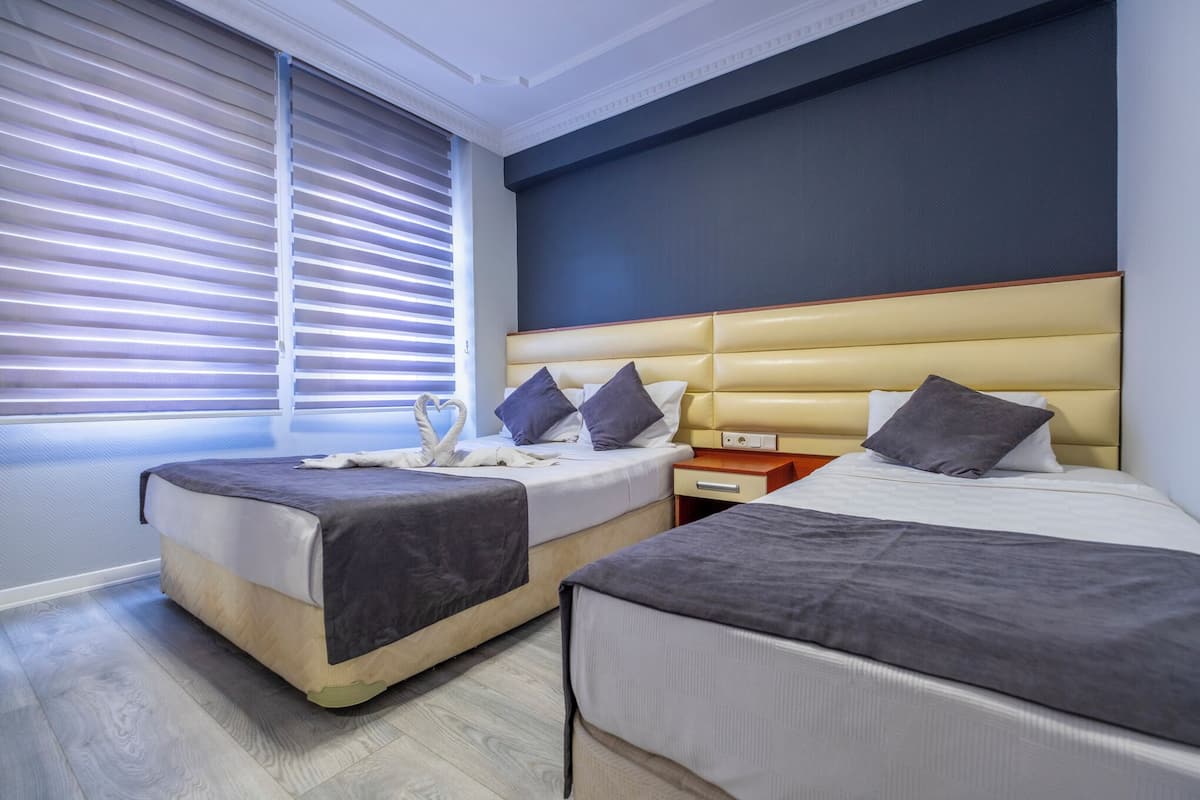 Une chambre d’hôtel moderne avec deux lits, une grande fenêtre avec stores, une tête de lit dotée de rangements et un plancher en bois.