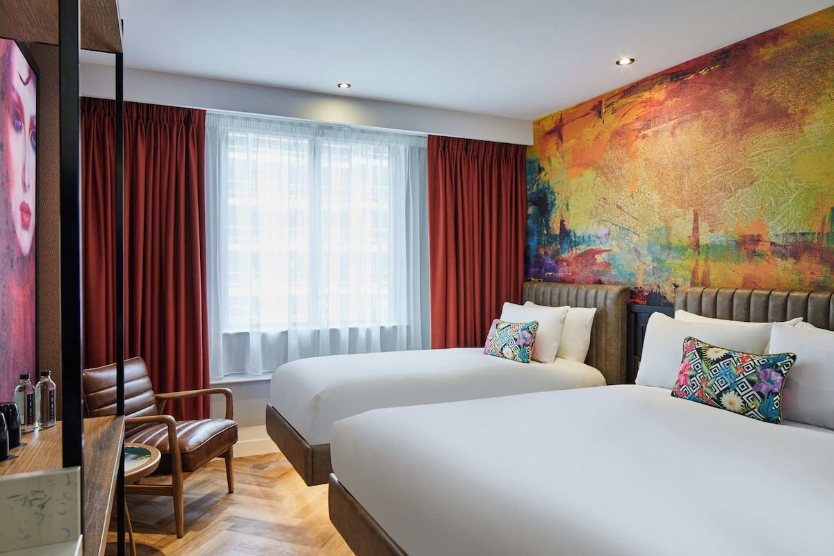 A hotel room with two beds, a desk with a chair, a large window with curtains, and a colorful abstract painting on the wall.