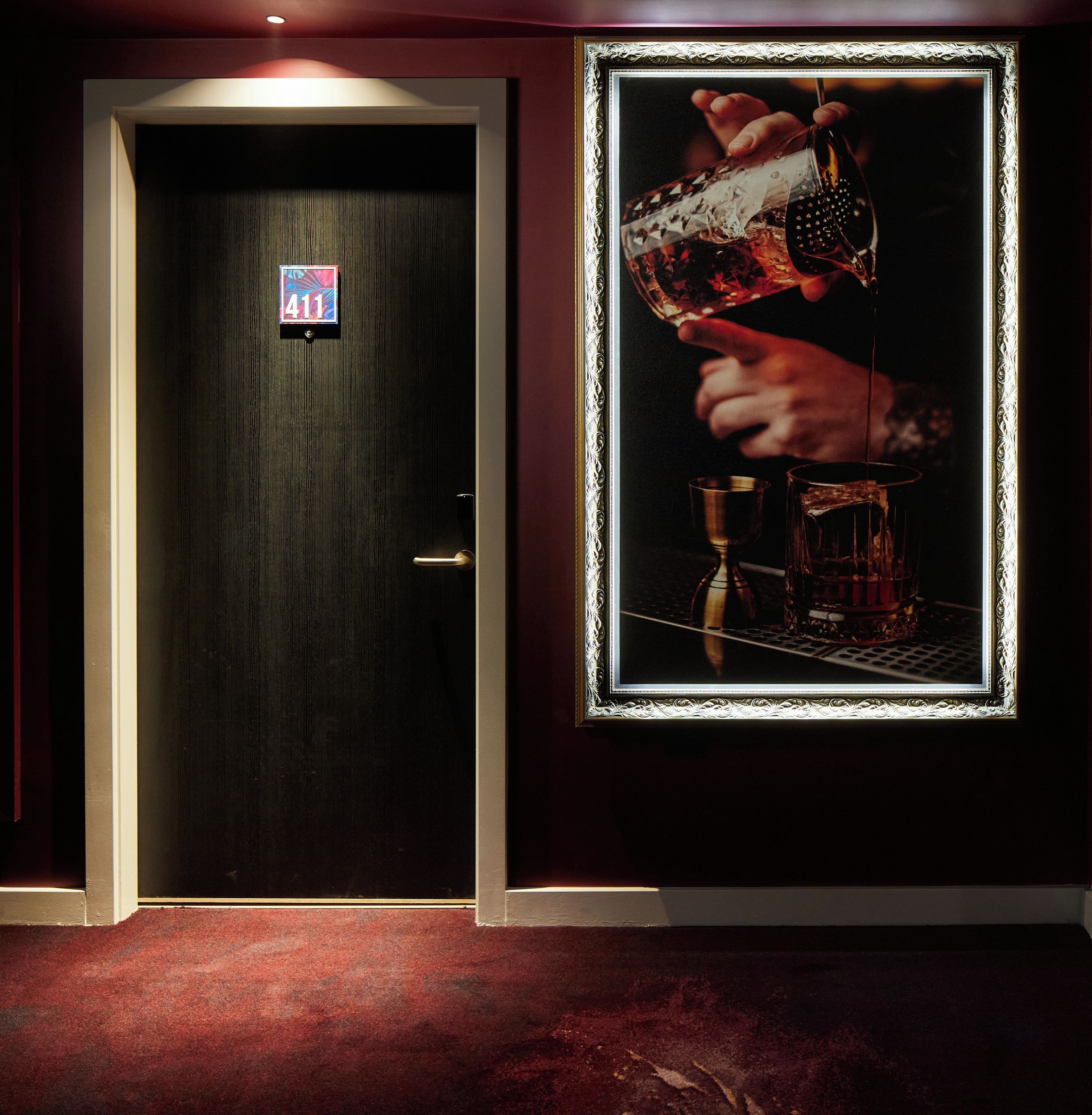 A door with a number sign, a framed picture of a hand pouring a drink, and a red carpeted floor.