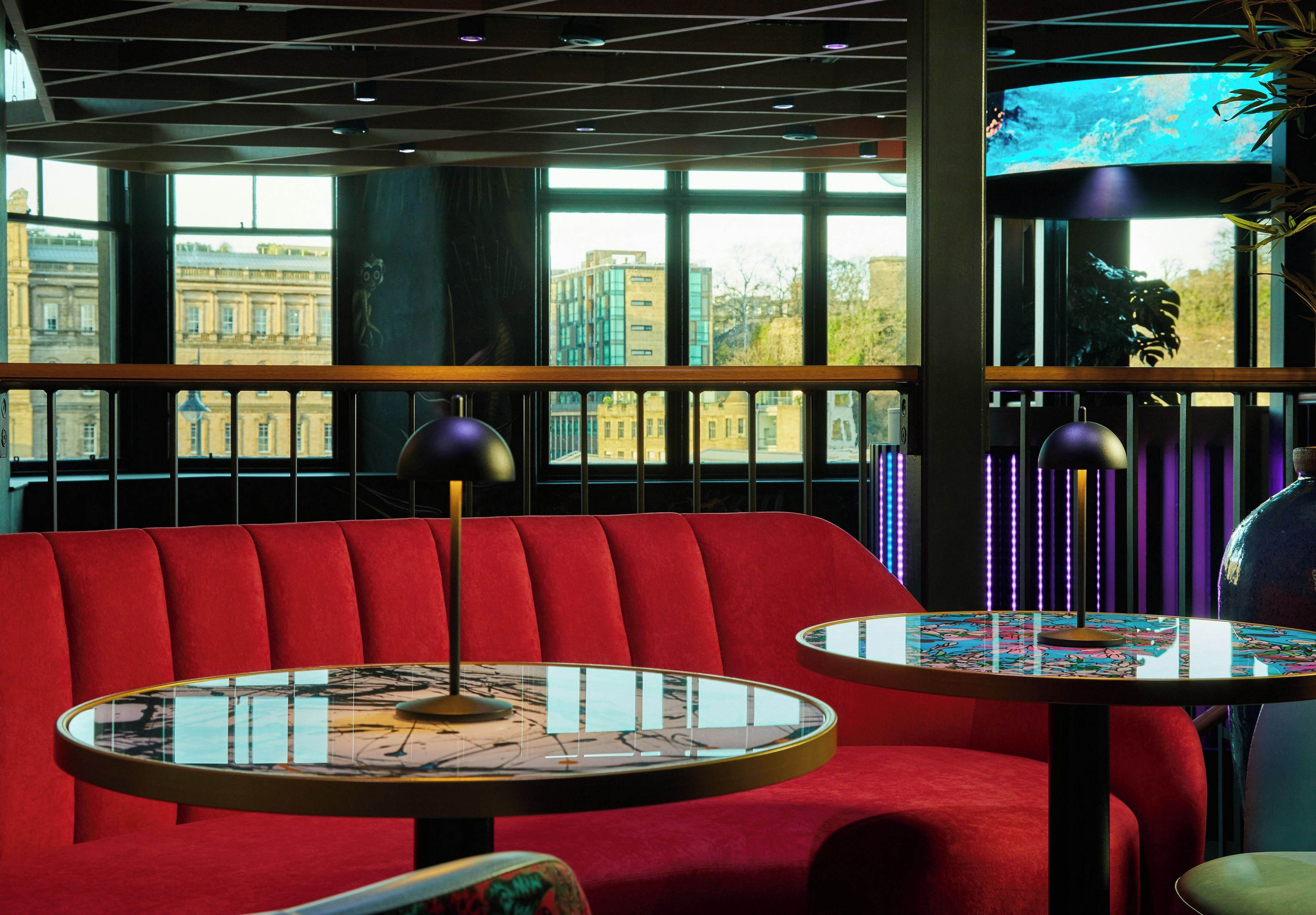 A modern interior with a red upholstered bench, two round tables with glass tops, and a city view through large windows.