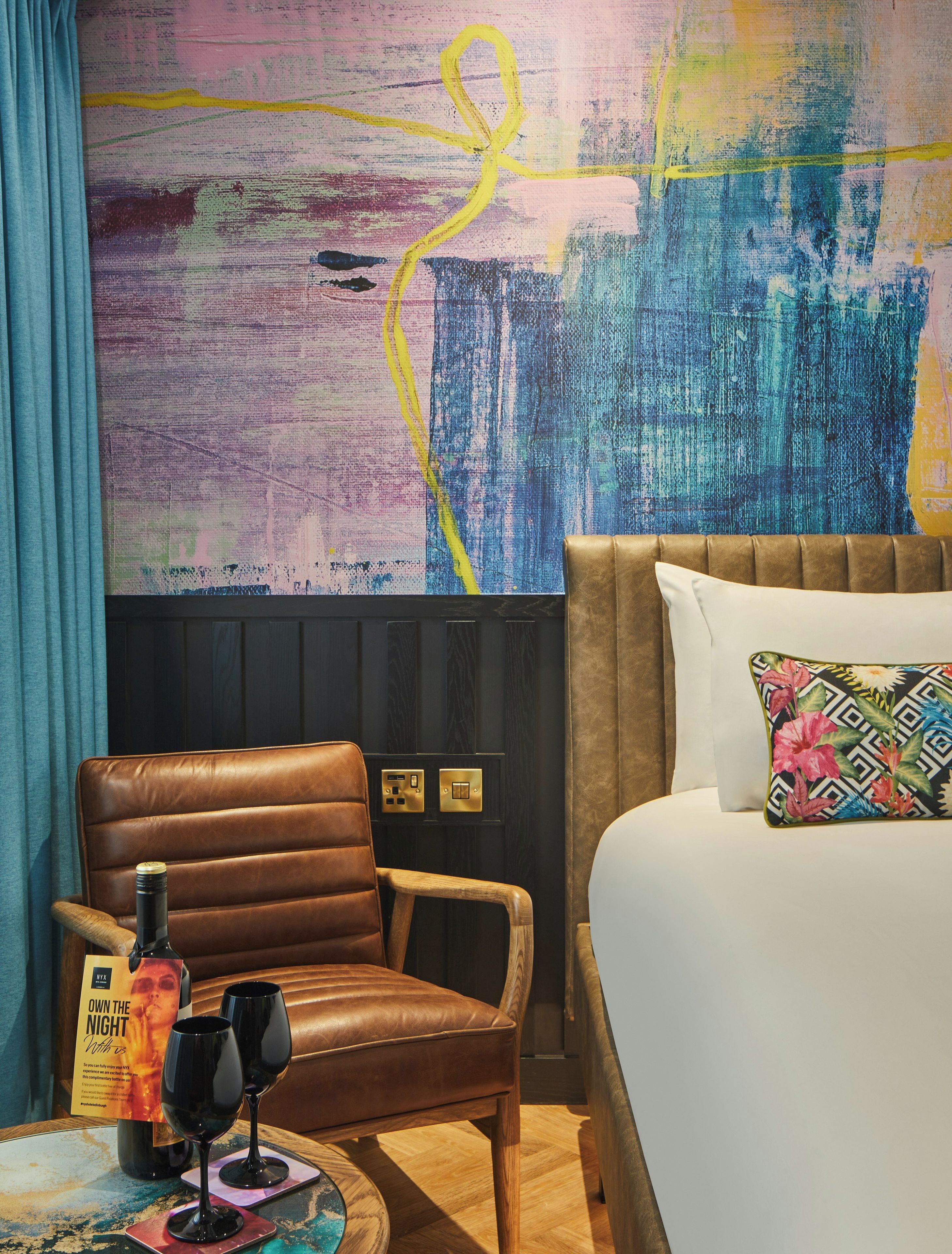 A hotel room with a brown leather chair, a bed with a colorful pillow, a wall with abstract art, and a bottle of wine on a coaster.