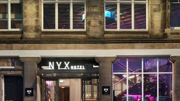 Entrada de un hotel con un letrero que dice 'NYX HOTEL', una puerta negra y un amplio ventanal con iluminación interior.
