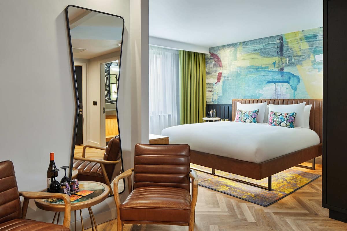 A hotel room with a large bed, a sitting area with a chair and table, a large mirror, and a colorful abstract painting on the wall.
