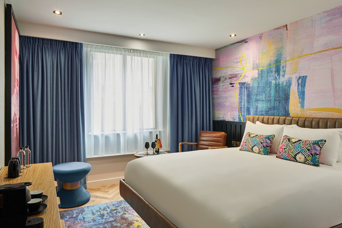 A modern hotel room with a large bed, a desk with a chair, a small table with two wine glasses, and a colorful abstract painting on the wall.