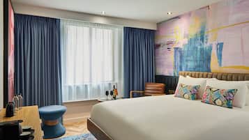 A modern hotel room with a large bed, a desk with a chair, a small table with two wine glasses, and a colorful abstract painting on the wall.