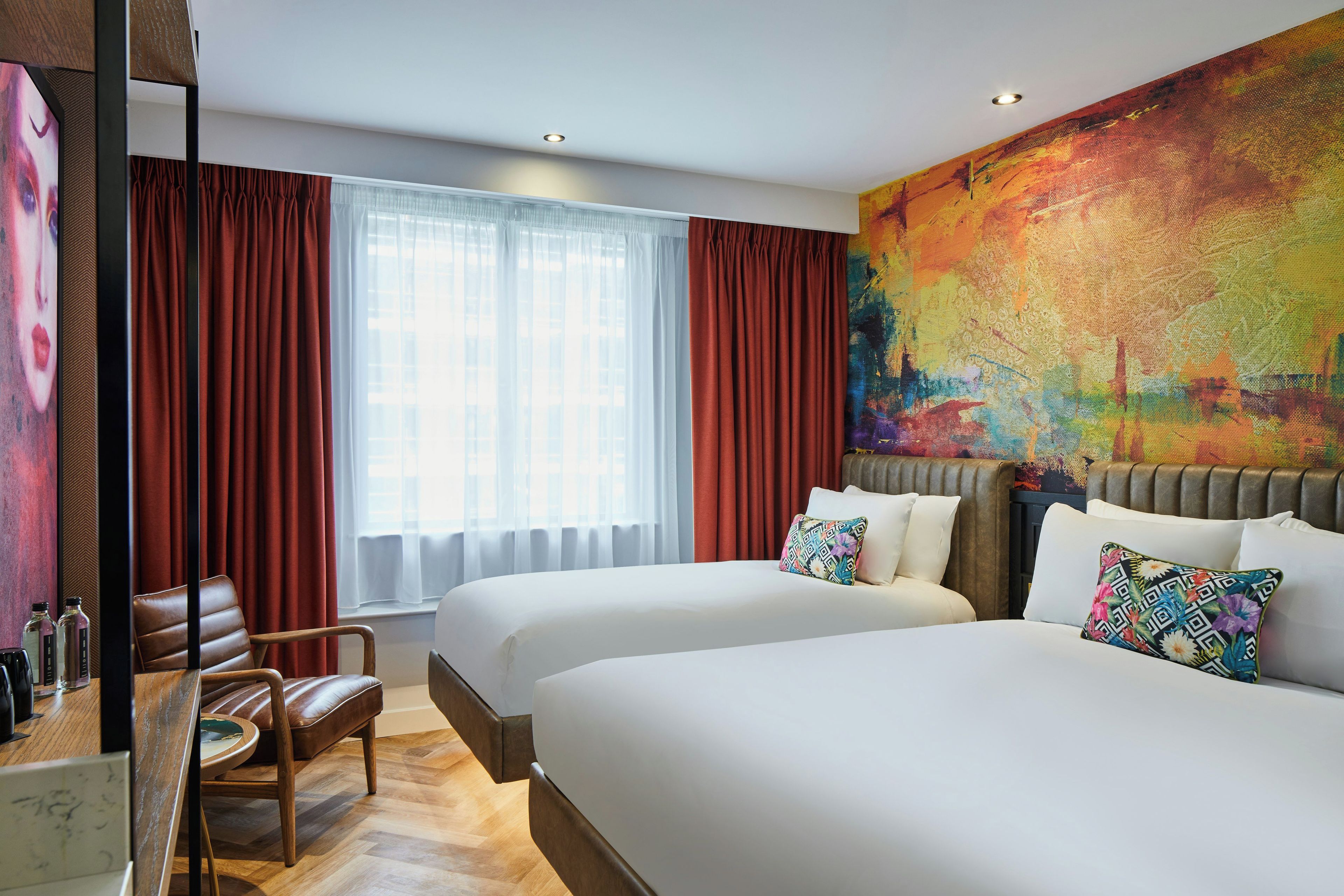 Hotellihuone, jossa on kaksi sänkyä, suuri ikkuna läpikuultavilla verhoilla, värikäs abstrakti maalaus seinällä ja puinen tuoli.