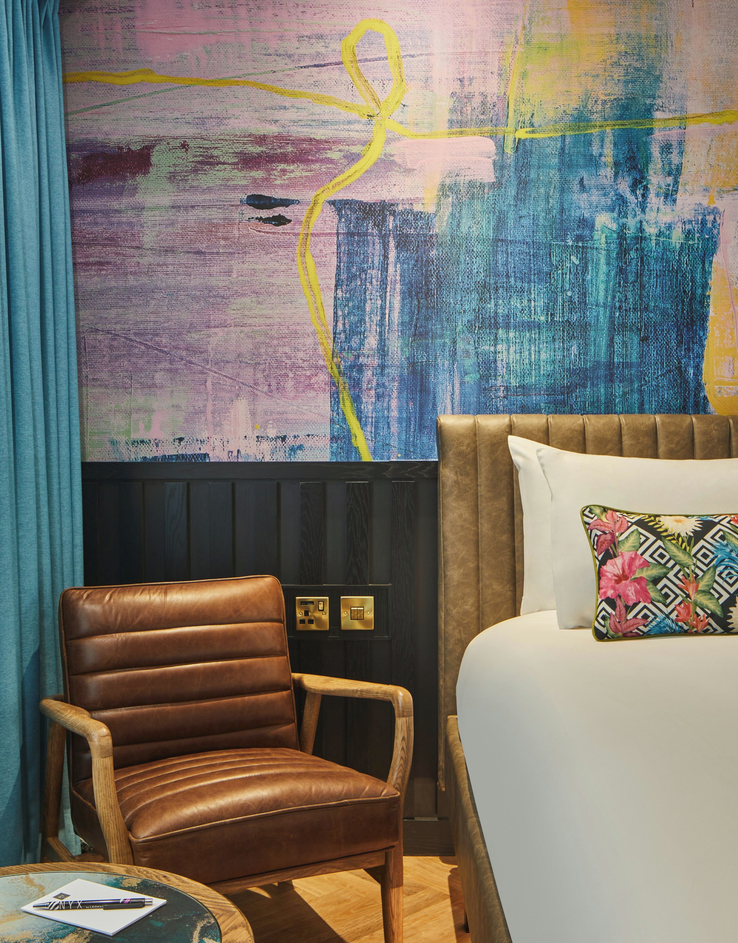 A hotel room with a brown leather chair, a bed with a patterned pillow, and a colorful abstract painting on the wall.