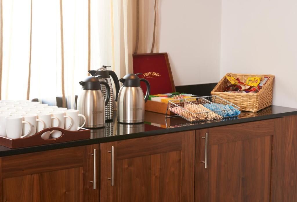 Una encimera de cocina con cafeteras, una bandeja de tazas blancas y una cesta de aperitivos.