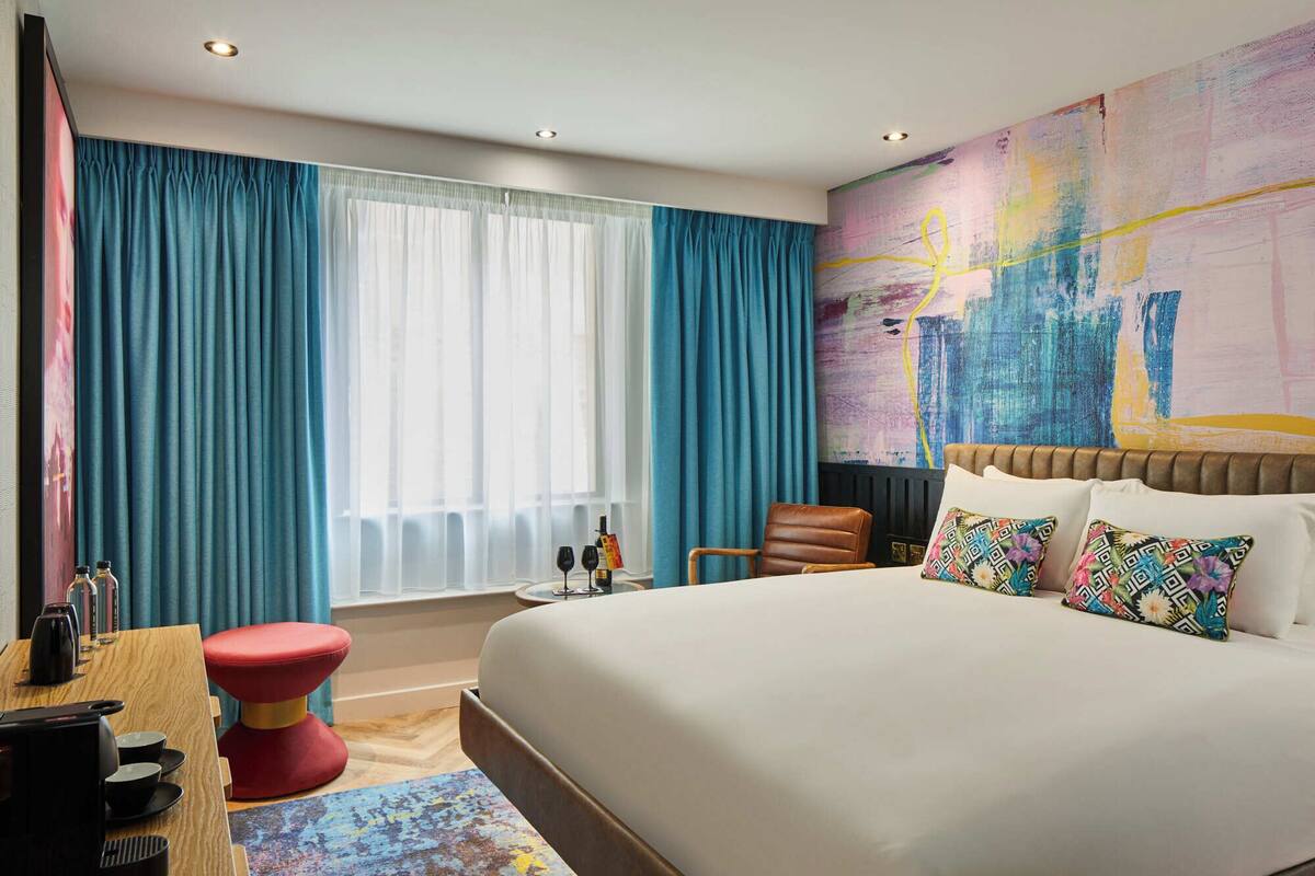 A modern hotel room with a large bed, a desk with a computer and coffee maker, a red stool, and a window with blue curtains.